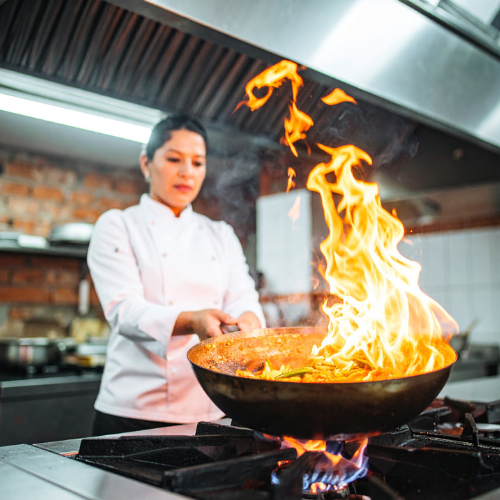 Professional female chef flambéing a dish in a sauté pan with flames rising over a gas stove in a modern commercial restaurant kitchen, showcasing live cooking and culinary expertise